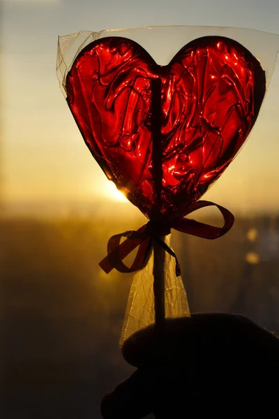 Red Lollipop in the form of heart in cellophane packaging with a ribbon on sunset sky background. Heart shaped lollipop. happy Valentines day idea. 14th february.