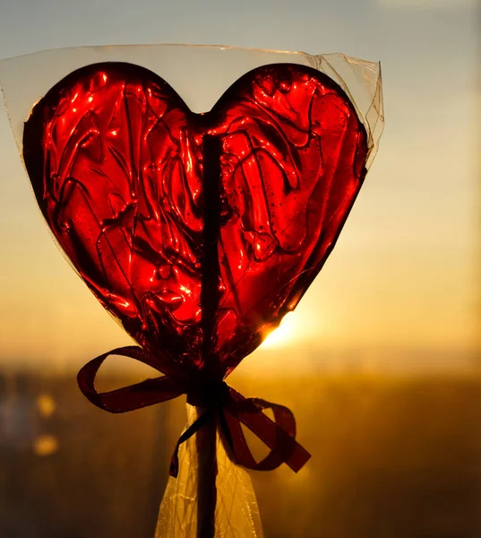 Red Lollipop in the form of heart in cellophane packaging with a ribbon on sunset sky background. Heart shaped lollipop. happy Valentines day idea. 14th february.