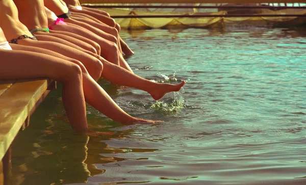 Group of women sitting in a row at the pool side. no faces. Group of girls sitting on a wooden platform by the river and splashing their feet in water on warm summer day. summertime season. beach party.