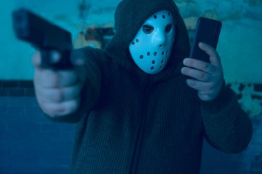 Kapüşonlu soyguncu eski fayanslı duvarlı arka planda silahı var. Adam beyaz hokey maskesi ve sıcak kazak giyiyor. Erkek, cep telefonunu tutuyor ve ekrana bakıyor..