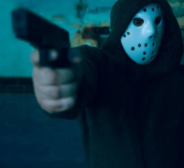 hooded robber with a gun on old tiled wall background. man wear white hockey mask and warm pullover.