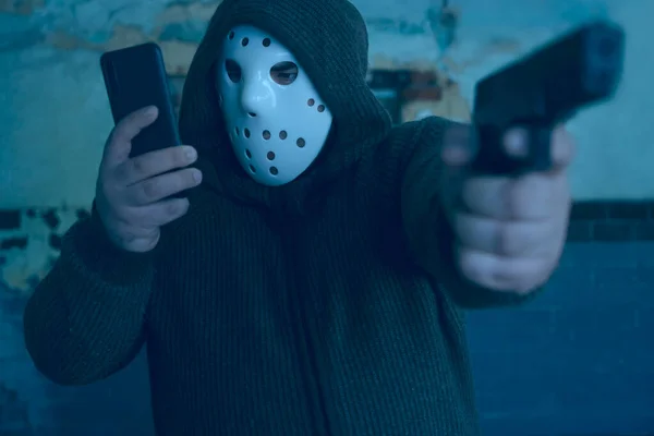 hooded robber with a gun on old tiled wall background. man wear white hockey mask and warm pullover. male holding in hand mobile phone and looking at screen.