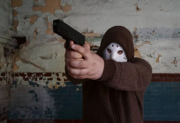 hooded robber with a gun on old tiled wall background. man wear white hockey mask and warm pullover.