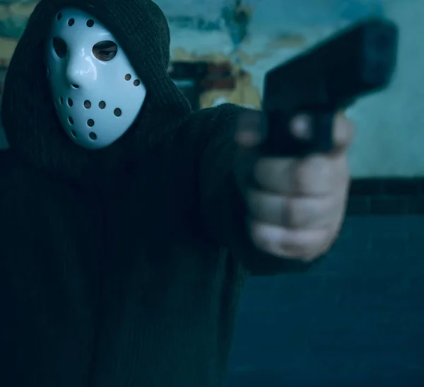 hooded robber with a gun on old tiled wall background. man wear white hockey mask and warm pullover.