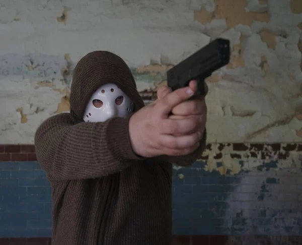 hooded robber with a gun on old tiled wall background. man wear white hockey mask and warm pullover.