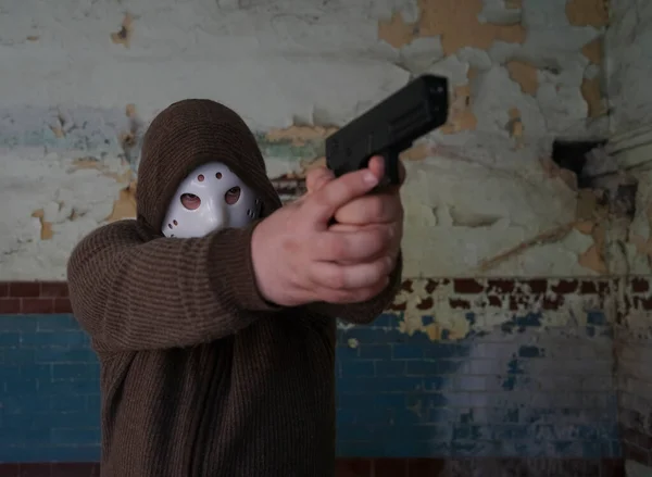 hooded robber with a gun on old tiled wall background. man wear white hockey mask and warm pullover.