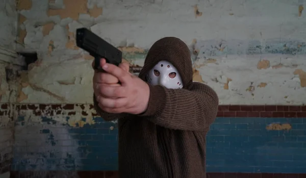 hooded robber with a gun on old tiled wall background. man wear white hockey mask and warm pullover.