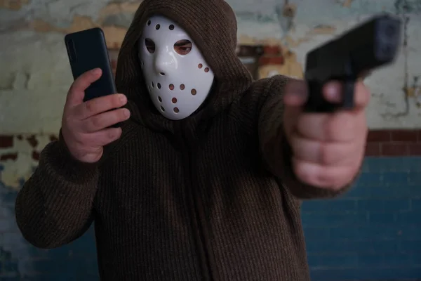 hooded robber with a gun on old tiled wall background. man wear white hockey mask and warm pullover. male holding in hand mobile phone and looking at screen.