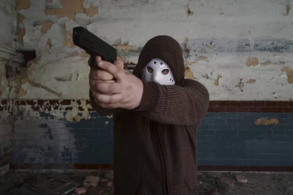 hooded robber with a gun on old tiled wall background. man wear white hockey mask and warm pullover.