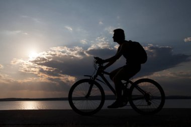 Bisikletli bir turistin gün batımına karşı gökyüzü bulutlu ve güneş ışınlı görüntüsü. Yalnız kadın. Siluet yarışı yüksek hızda