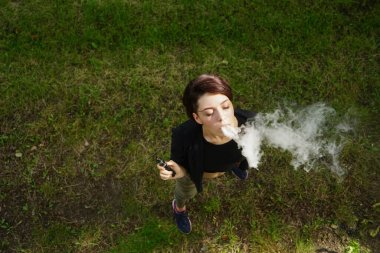 Genç kız elektronik sigara içiyor. Yeşil çimen arka planında buharlaştırıcı buharlaştıran bir genç. Buharlaşan genç bir kadının yüzü. Üst Manzara. Sigara günü yok.