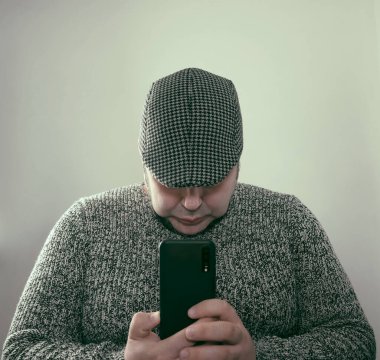 handsome man wearing stylish white and black sweater and cap.  isolated on gray background. no eyes. unrecognizable person. male looking in screen of mobile phone. 