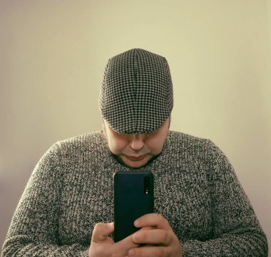 handsome man wearing stylish white and black sweater and cap.  isolated on gray background. no eyes. unrecognizable person. male looking in screen of mobile phone. 