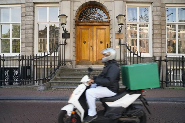 Food delivery moto scooter driver with white backpack behind back is on his way to deliver food. Courier on scooter delivering food. Quick shipping of goods to customers. Amsterdam, Netherlands
