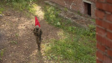 Modern kamuflajlı asker terk edilmiş binanın yakınında keskin nişancı tüfeğiyle yürüyor. Adam kırmızı bayrak taşıyor..  