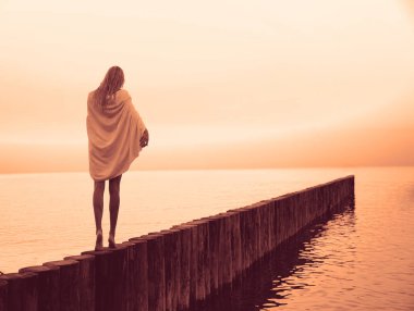 Kafkasyalı sarışın kız omuzlarında beyaz ıslak havluyla gün batımı kumsalında yürüyor.. 