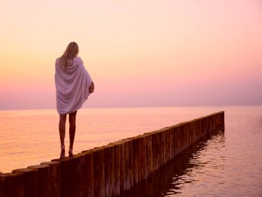 Kafkasyalı sarışın kız omuzlarında beyaz ıslak havluyla gün batımı kumsalında yürüyor.. 