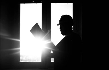 Bir erkek mühendis, güneşli pencerenin arka planında bir kağıt planıyla ayakta duruyor.. 