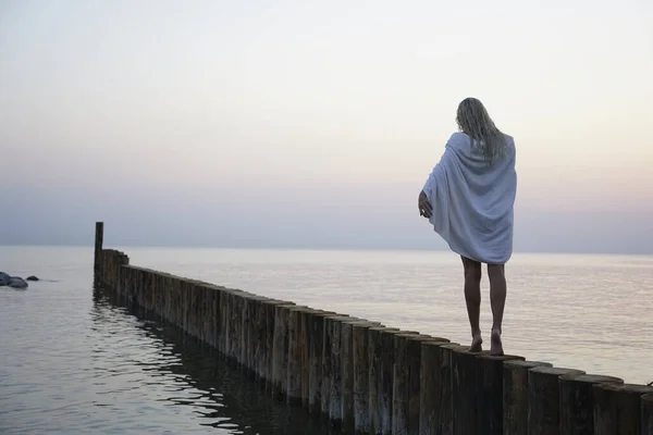 caucasian blonde girl walking on sunset beach with  white wet towel on shoulders.