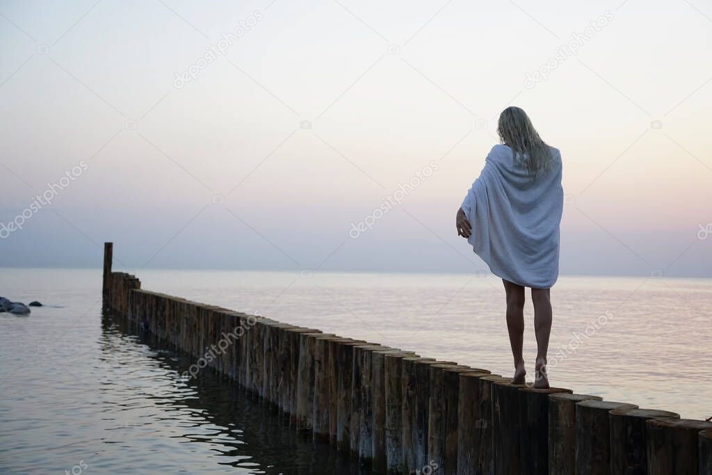 caucasian blonde girl walking on sunset beach with  white wet towel on shoulders. 