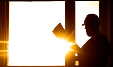 Bir erkek mühendis, güneşli pencerenin arka planında bir kağıt planıyla ayakta duruyor.. 