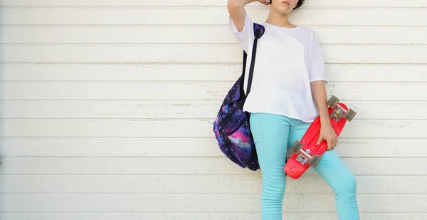 street style  woman in white t-shirt and colorful trousers with skateboard standing outdoors. garage doors on background. full length.  summertime season.