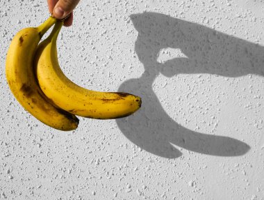 Erkek elinde sarı muzlar. Arka planda beyaz beton duvar var. Çitin üzerinde güneş ışığı ve gölge. 