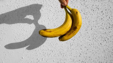 Erkek elinde sarı muzlar. Arka planda beyaz beton duvar var. Çitin üzerinde güneş ışığı ve gölge. 