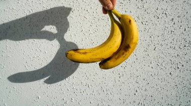 Erkek elinde sarı muzlar. Arka planda beyaz beton duvar var. Çitin üzerinde güneş ışığı ve gölge. 