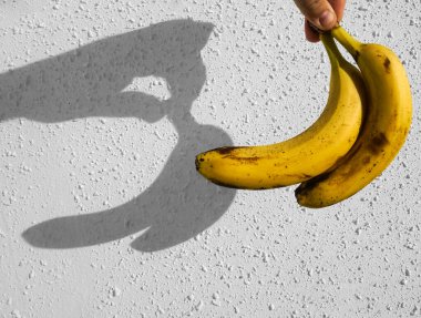 Erkek elinde sarı muzlar. Arka planda beyaz beton duvar var. Çitin üzerinde güneş ışığı ve gölge. 