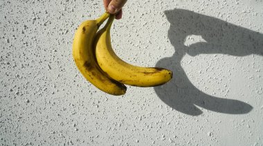 Erkek elinde sarı muzlar. Arka planda beyaz beton duvar var. Çitin üzerinde güneş ışığı ve gölge. 