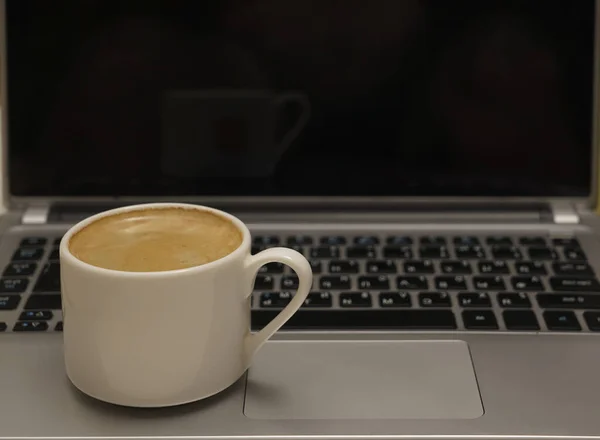 White Cup of coffee with pattern on the foam. Hot freshly brewed coffee in a white mug on gray laptop. empty black screen.