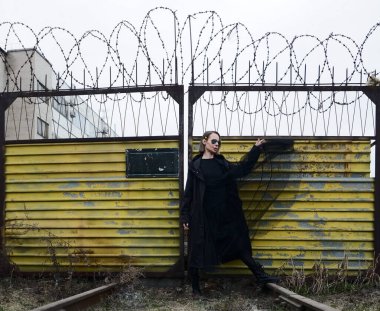 Sarı saçlı genç bir kız, kapalı sarı metal bir kapının yanında duruyor. Siyah elbiseli kadın..