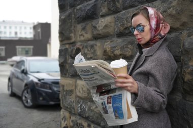 MOSCOW, RUSSIA - 27 Nisan 2021: şık şık bir kadın sıcak kahve fincanı siparişiyle ayakta duruyor ve taş duvar arkasında gazete okuyor.