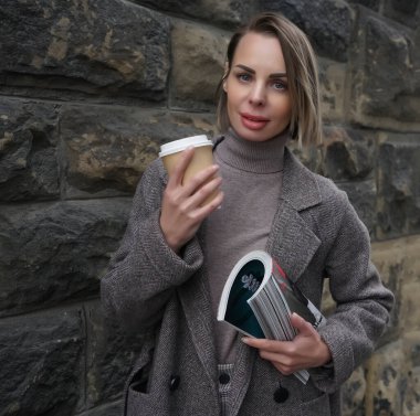 MOSCOW, RUSSIA - 27 Nisan 2021: orta yaşlı, gri ceketli, elinde moda dergisi ve kahve fincanı tutan kadın. Uzaklara bakan bir kadın.