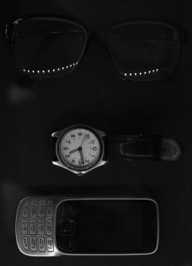 Gözlükler, eski model cep telefonu ve siyah masa örtüsünün üzerinde uzanan bir saat. Siyah ve beyaz kompozisyon. Düz yatıyordu.
