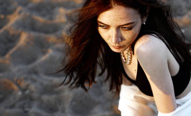 Kızıl saçlı, beyaz tişörtlü, gün batımının arka planında kumların üzerinde şehvetli bir kız.. 