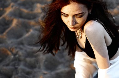 Kızıl saçlı, beyaz tişörtlü, gün batımının arka planında kumların üzerinde şehvetli bir kız.. 