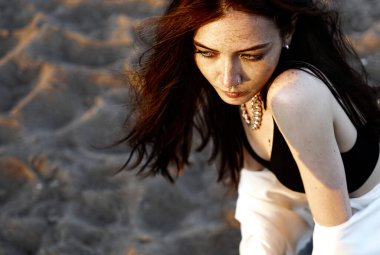 Kızıl saçlı, beyaz tişörtlü, gün batımının arka planında kumların üzerinde şehvetli bir kız.. 