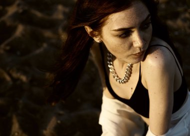 Beyaz tişörtlü kızıl saçlı kız gün batımı kumsalının arka planında kumları düşünüyor. Yaz mevsimi. 