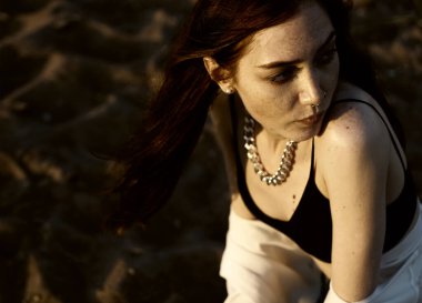 Beyaz tişörtlü kızıl saçlı kız gün batımı kumsalının arka planında kumları düşünüyor. Yaz mevsimi. 