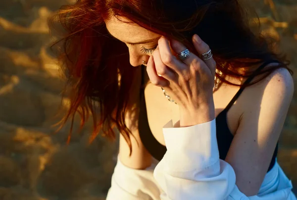 白いシャツを着た赤毛の官能的な女の子が夕日のビーチの背景に砂の上に座って考えている 夏の季節 ブラック 指には銀の指輪 — ストック写真