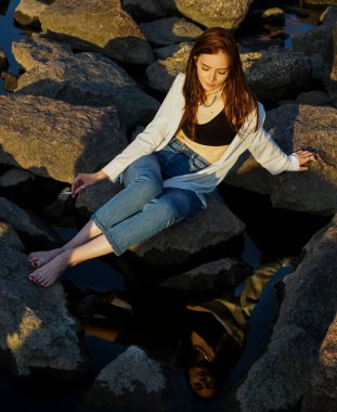 Kız elinde sigarayla koca bir taşın üzerinde oturuyor. Sudaki yansıma