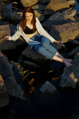 Kız elinde sigarayla koca bir taşın üzerinde oturuyor. Sudaki yansıma