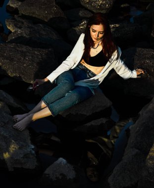 Kız elinde sigarayla koca bir taşın üzerinde oturuyor. Sudaki yansıma