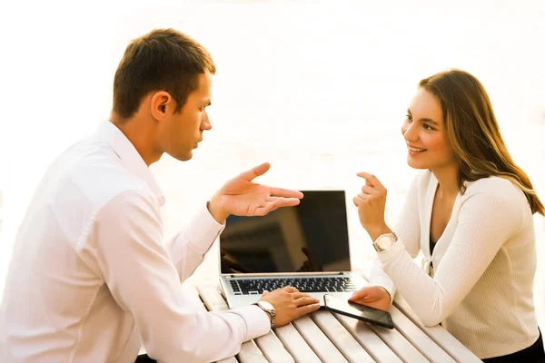 two business people  at coffee table while in a meeting outdoors, using a laptop and cell phone.