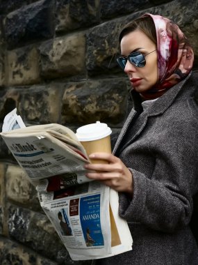 MOSCOW, RUSSIA - 27 Nisan 2021: şık şık bir kadın sıcak kahve fincanı siparişiyle ayakta duruyor ve taş duvar arkasında gazete okuyor.
