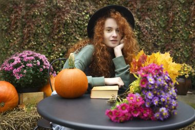 Sıcak bir atmosferde romantik bir kitap okuyan genç bir kadın hayal ediyorum. Kızıl saçlı bayan sonbahar havasının tadını çıkarıyor. Sarı akçaağaç çiçekli ve turuncu kabaklı siyah masa.. 