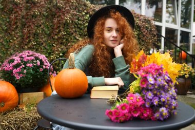Sıcak bir atmosferde romantik bir kitap okuyan genç bir kadın hayal ediyorum. Kızıl saçlı bayan sonbahar havasının tadını çıkarıyor. Sarı akçaağaç çiçekli ve turuncu kabaklı siyah masa.. 