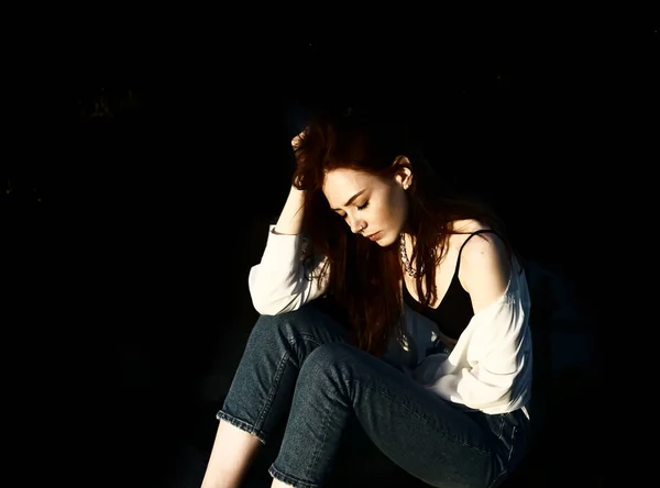 長い赤い髪のジーンズを着た若い美しい女性が床に座っています 目を閉じて 黒い背景に隔離され — ストック写真